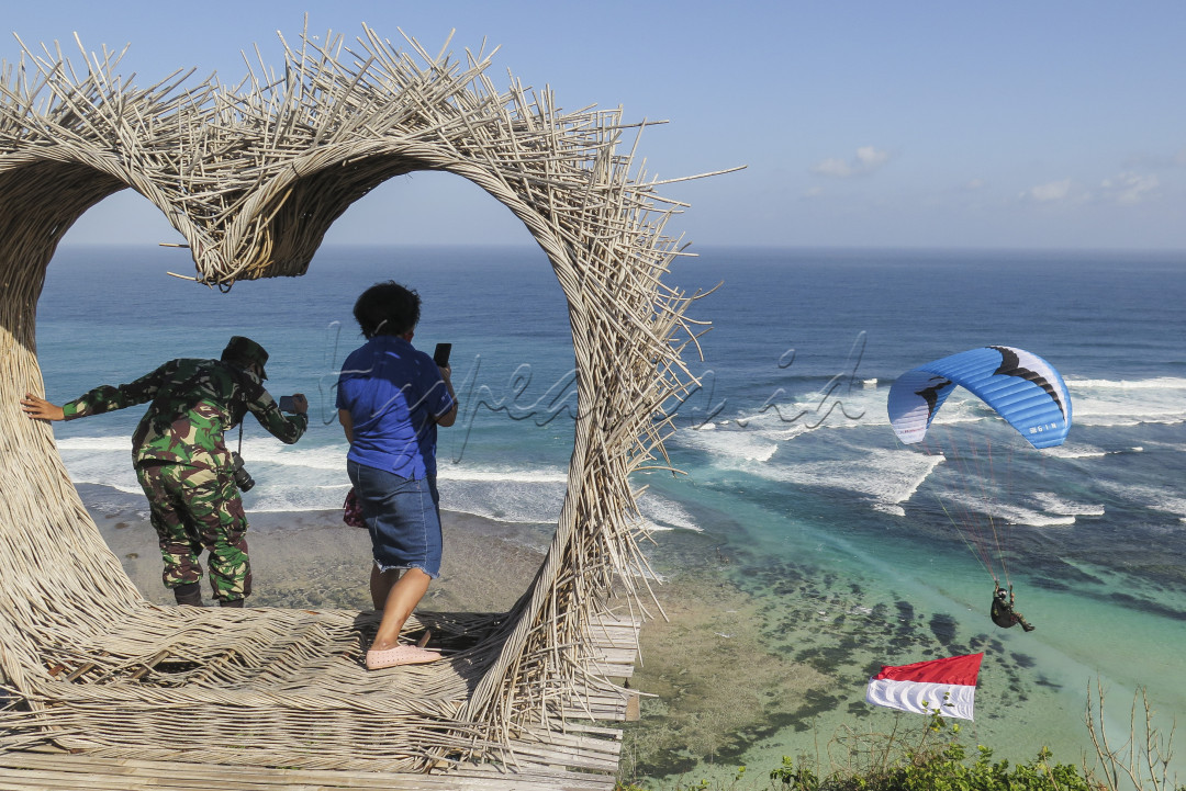Anggota TNI bersama warga mendokumentasikan seorang anggota TNI lainya yang sedang terbang Paralayang dengan membawa bendera Merah Putih diatas pantai bukit Timbis, Nusa Dua, Bali pada 7 Juli 2020. Kegiatan tersebut merupakan rangkaian latihan bersama jelang perayaan Hari Kemerdekaan Indonesia Ke-75.&nbsp;