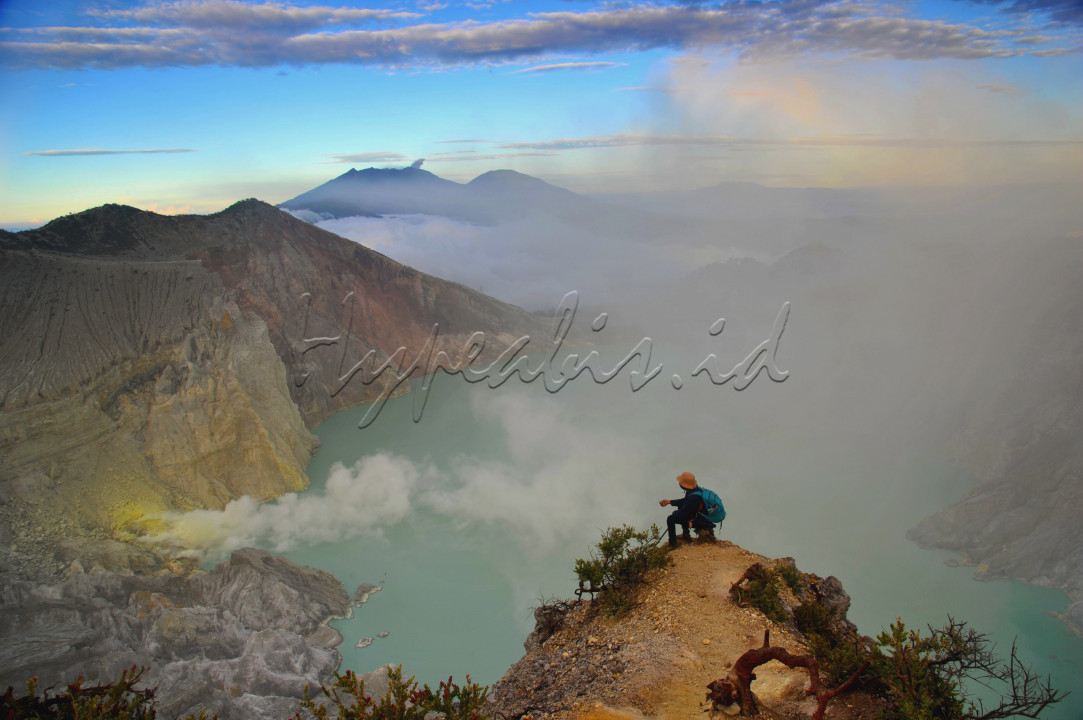 Seorang pendaki gunung sedang menikmati pesona Kawah Ijen saat menjelang matahari terbit. Kawah Ijen dari atas puncak Gunung Ijen terlihat sangat indah dengan danau besar berwarna hijau kebiruan serta kabut dan asap belerang yang sangat memesona. Gunung Kawah Ijen atau Kawah Ijen terletak di antara Kabupaten Bondowoso dan Kabupaten Banyuwangi yang merupakan salah satu gunung aktif di Jawa Timur.&nbsp;