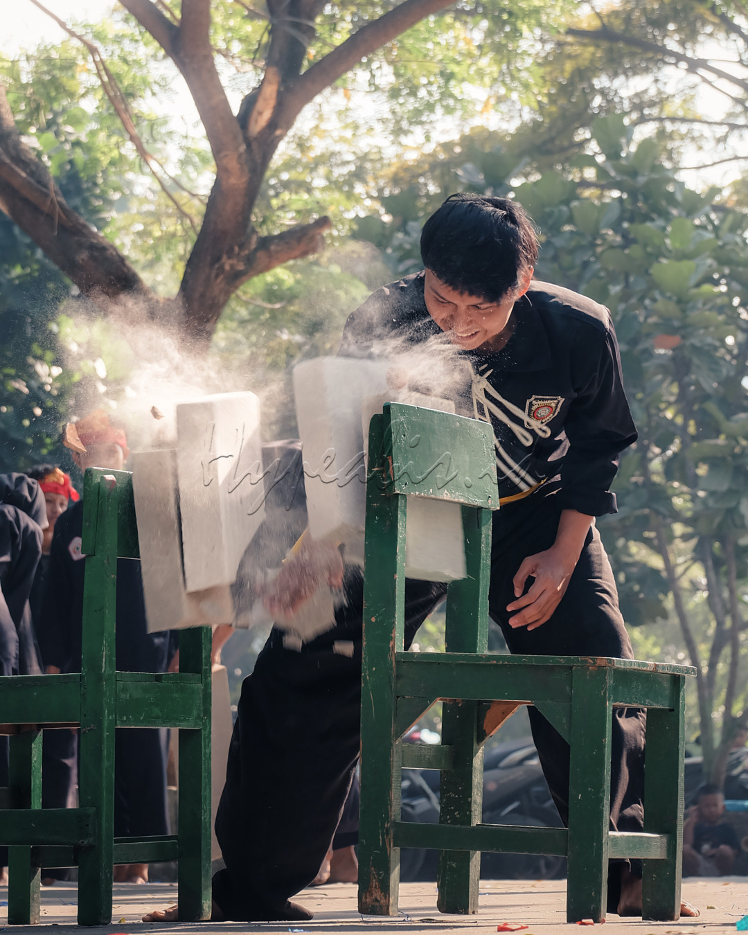 Dalam foto ini, seorang praktisi bela diri sedang menunjukkan kekuatan dan keterampilannya dengan memecahkan batu menggunakan tangannya. Aksi ini dilakukan di luar ruangan, di bawah naungan pepohonan, dan disaksikan oleh penonton. Debu dan pecahan batu beterbangan di udara, menambah dramatisasi momen ini. Ini adalah contoh dari demonstrasi kekuatan dan ketangguhan yang sering ditampilkan dalam pertunjukan bela diri.