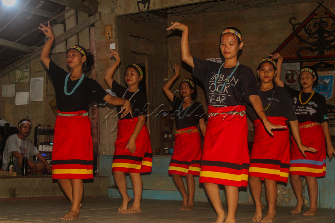 Foto ini memperlihatkan tarian khas Mentawai yang berlangsung di malam hari dalam uma, rumah adat Mentawai. Para penari dengan gerakan yang penuh makna, menari di sebuah rumah adat Mentawai, membawa keajaiban dan kekayaan budaya ke dalam suasana malam. Tarian ini mencerminkan hubungan yang mendalam antara tradisi dan kehidupan sehari-hari suku Mentawai, yang terus dipelihara dengan penuh kebanggaan.