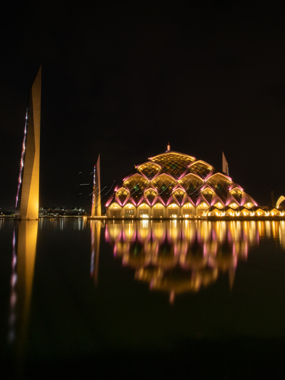 Masjid Al-Jabbar di Bandung saat malam hari menampilkan pemandangan yang memukau. Masjid ini memiliki desain arsitektur modern dengan atap berbentuk seperti prisma dan dikelilingi oleh kolam air yang mencerminkan cahaya lampu, menciptakan efek visual yang menakjubkan. Pada malam hari, masjid ini diterangi oleh lampu-lampu warna-warni yang memancarkan cahaya keemasan dan biru, menambah kesan megah dan sakral. Pemandangan ini semakin indah dengan latar belakang langit malam yang gelap, menciptakan suasana yang damai dan spiritual.