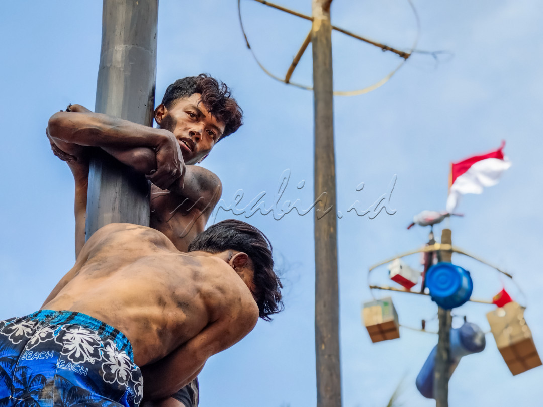 Dua pemuda berjuang memanjat tiang licin dalam tradisi Panjat Pinang, sebuah lomba yang ikonik dan penuh semangat di perayaan Hari Kemerdekaan Indonesia. Bendera Merah Putih berkibar di latar belakang, melambangkan kebanggaan dan persatuan bangsa.