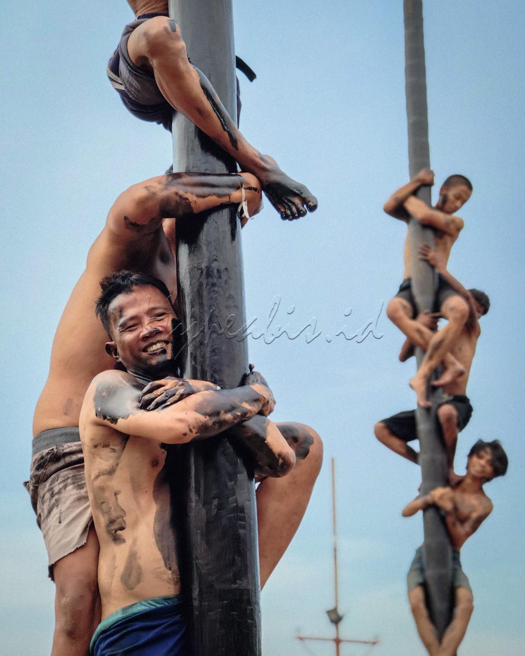 Dalam foto ini tergambar semangat kebersamaan yang kuat, tercermin dalam tradisi panjat pinang, sebuah lomba yang kerap diselenggarakan saat perayaan Hari Kemerdekaan Indonesia. Para peserta, dengan penuh tawa dan usaha, saling bahu-membahu memanjat batang pinang yang licin demi mencapai puncak. Ekspresi wajah mereka tidak hanya menggambarkan keceriaan, tetapi juga tekad dan kerja keras yang menjadi cerminan karakter bangsa Indonesia. Foto ini menangkap esensi dari "Wajah Indonesia" yang penuh dengan semangat gotong royong, kebersamaan, dan keceriaan.