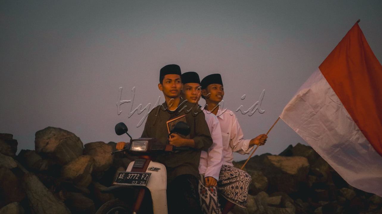Tiga santri sedang mengendarai sepeda motor bersama, mereka mengenakan pakaian tradisional santri. salah satunya&nbsp;memegang bendera merah putih dengan bangga, yang berkibar tertiup angin saat mereka melaju.Bendera ini terlihat mencolok dan menjadi fokus utama foto, melambangkan kebanggaan mereka sebagai warga negara Indonesia.&nbsp;Ekspresi yang penuh semangat dan tekad menambahkan kesan keteguhan mereka dalam beriman dan bangga terhadap negara.&nbsp;mencerminkan perpaduan antara semangat keagamaan dan nasionalisme, menunjukkan bagaimana mereka dengan tegas melanjutkan perjalanan mereka sambil mengibarkan bendera sebagai simbol identitas dan kebanggaan mereka.