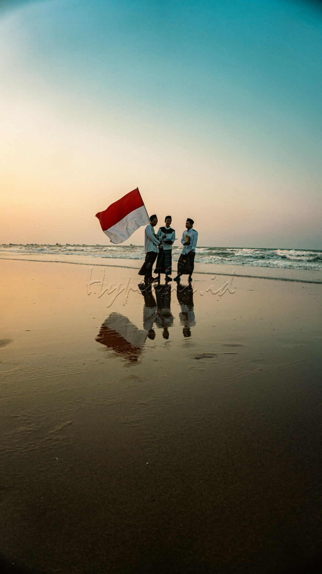 Momen yang penuh simbolisme dan keindahan.&nbsp;tampak tiga orang santri yang berdiri di pesisir pantai, dengan latar belakang pantai yang luas dan laut yang biru berkilauan.&nbsp;Refleksi mereka di air menciptakan efek visual yang dramatis, menambahkan dimensi dan kedalaman pada gambar. Bendera merah putih tampak berkibar dengan lembut di tangan mereka, dan pantulannya di air memperkuat kesan patriotisme dan kekuatan simbolis dari bendera tersebut. Masing masing dari mereka membawa kitab, mendedikasikan mereka pada ciri khasnya yaitu belajar. Seolah olah mereka menggabungkan kecintaan terhadap negara dan agama dalam satu momen harmonis.&nbsp;sebuah foto yang menyoroti perpaduan antara iman dan kebanggaan nasional dalam konteks alam yang menenangkan.