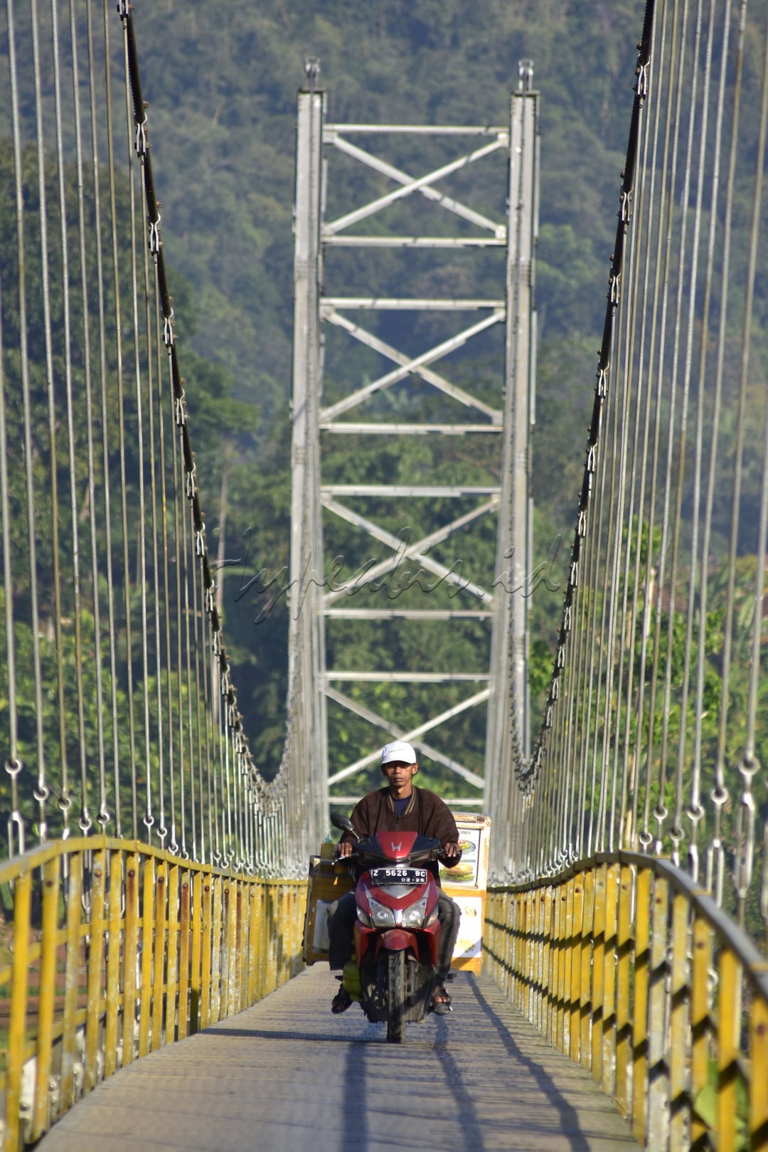 Seorang pedagang dengan motornya melintasi jembatan gantung untuk menjajakan dagangannya di desa seberang ,&nbsp;