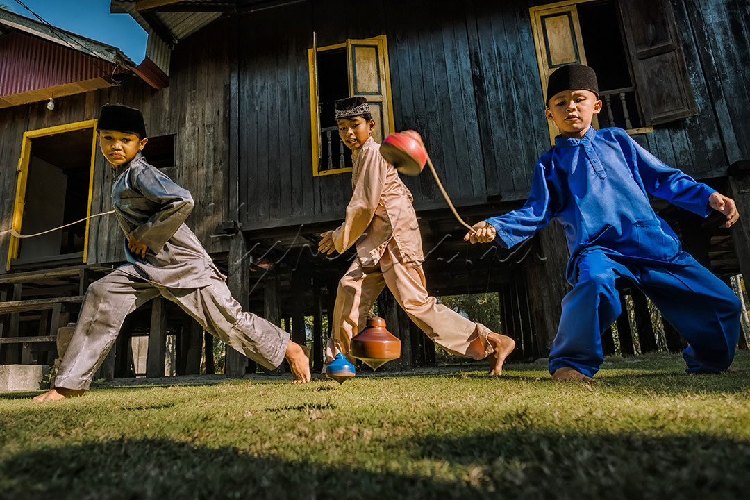Tiga orang anak mengadu ketangkasan memainkan gasing dengan latar belakang rumah melayu.<br />
Permainan gasing merupakan salah satu permainan tradisional Indonesia yang menjadi warisan budaya tak benda dari bumi melayu.&nbsp;<br />
&nbsp;