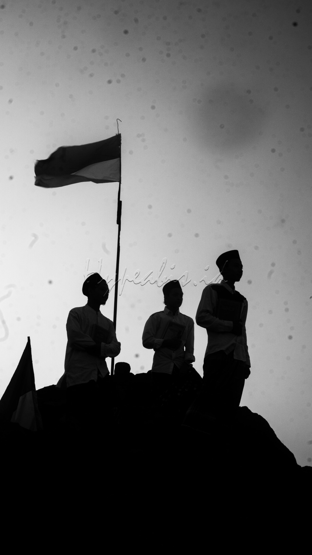 Tiga orang santri berdiri tegak di atas bebatuan besar di tepi pantai, mengenakan pakaian khas mereka. mereka memegang kitab di dadanya denga hormat.&nbsp;Bendera yang terpasang di belakang mereka, meskipun dalam warna hitam putih, tampak menonjol dengan jelas, berkibar lembut, memberikan efek dramatis yang sejalan dengan suasana serius dan penuh makna dari santri.