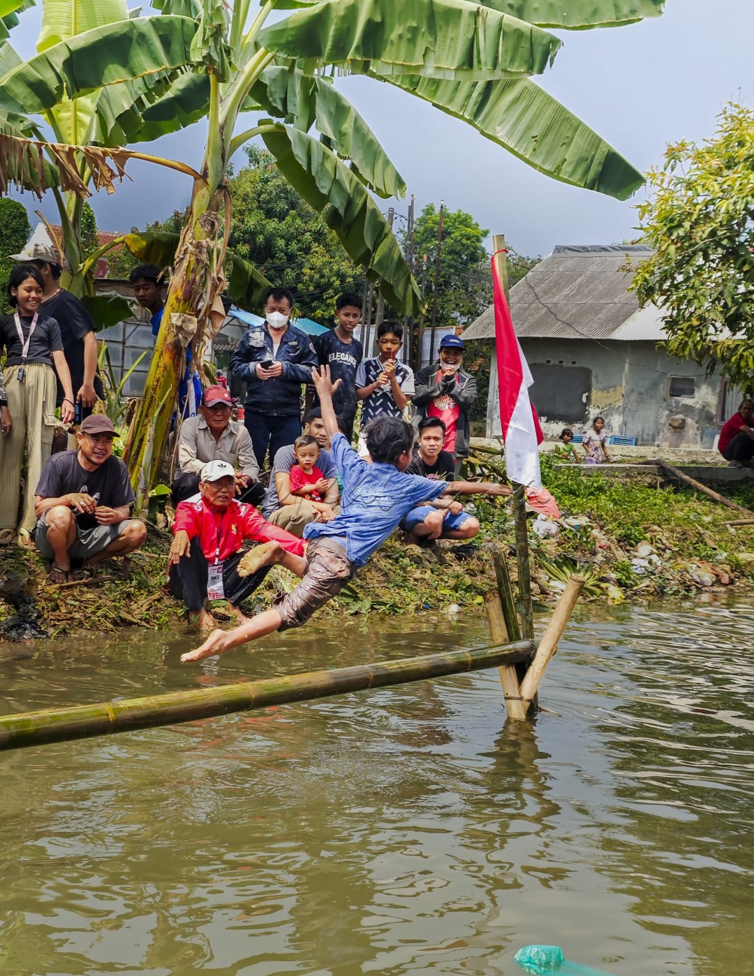 Seorang Anak yg sedsng mengikuti lomba keseimbangan melewati bambu diatas air, berhasil mencapai ujung dan meraih bendera merah putih sambil meloncat.