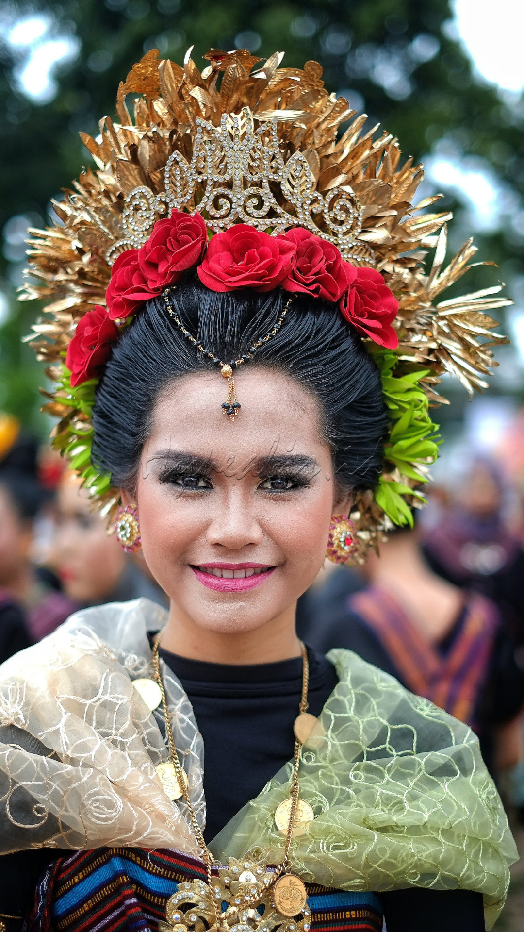 Seorang wanita dengan pakaian adat dan mahkota yang indah, tersenyum dengan ekspresi bahagia.