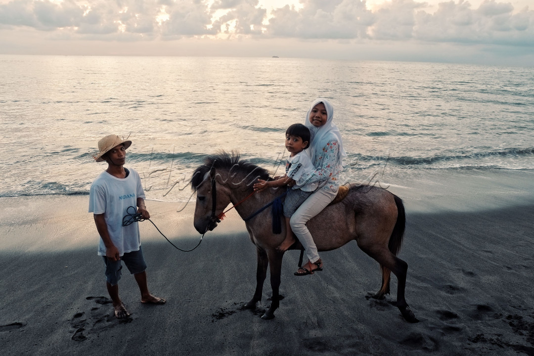Dua orang sedang mengendari kuda di tepi pantai yang indah dengan pemandangan laut yang luas di latar belakang.