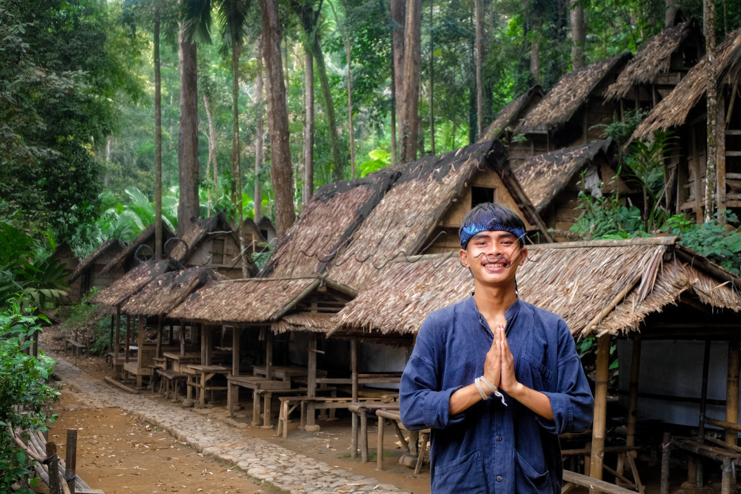 Damidi, adalah sosok pemuda Baduy Luar memberikan salam ramah dengan pakaian khas Baduy Luar yang berwarna biru tua dan hitam dengan latarbelakang barisan 'Leuit' atau lumbung pangan tradisional yang dikelilingi hutan yang masih dijaga kelestariannya. Foto diambil pada tanggal 15 Agustus 2024