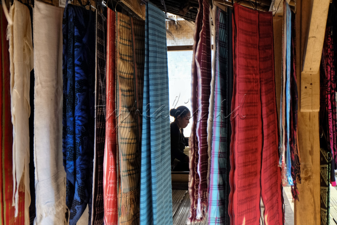 Seorang wanita suku Baduy Luar menenun kain tradisional Baduy yang penuh makna dan nilai historis, berlatar depan berbagai macam kain khas hasil tenunan. Selain menjaga nilai-nilai kebudayaan, kain tenun menjadi salah satu penggerak perekonomian masyarakat Baduy Luar. Foto diambil pada tanggal 15 Agustus 2024