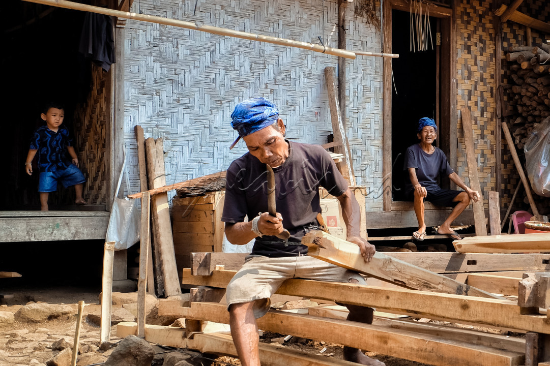 Seorang warga Baduy Luar sedang bekerja membuat pondasi rumah adat dengan latar belakang seorang anak dan orang yang lebih tua menyimbolkan kebudayaan yang dijaga dari generasi ke generasi. Foto diambil pada tanggal 15 Agustus 2024