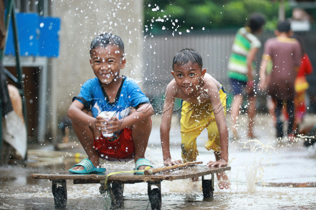 Sebuah tawa riang bergema di tengah hujan yang turun deras. Sejumlah anak-anak tampak asyik bermain di genangan air yang terbentuk di jalanan. Wajah mereka merefleksikan kegembiraan, tak peduli dengan rintikan hujan yang membasahi tubuh.<br />
Mereka berlarian, bercanda, dan saling menyiram dengan air. Seorang anak laki-laki bahkan melompat dengan lincah, membuat percikan air yang indah. Kepolosan dan keceriaan anak-anak ini mengingatkan kita akan betapa sederhana dan menyenangkannya bermain di tengah alam, bahkan di saat hujan.<br />
Hujan yang biasanya menjadi penghalang bagi sebagian orang, bagi mereka justru menjadi sumber kebahagiaan. Kesenangan bermain di tengah hujan menjadi momen yang tak terlupakan bagi mereka. Foto ini merekam momen indah tersebut, momen yang sarat dengan keceriaan dan kepolosan anak-anak yang sedang menikmati masa kanak-kanak.