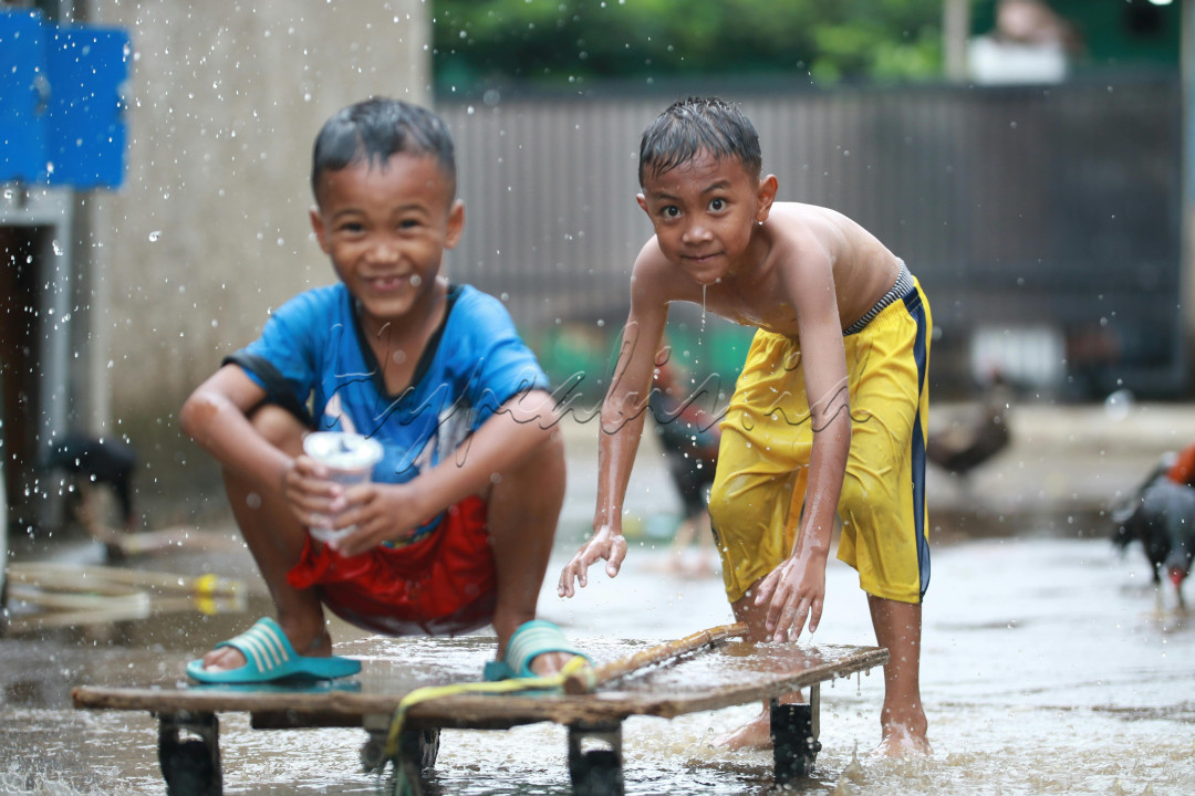 Dua anak laki-laki bermain dengan riang di tengah hujan deras. Anak yang berada di sebelah kanan tampak berlari dengan penuh semangat sambil mengacungkan tangan kanannya ke arah kamera. Ekspresi wajahnya menggambarkan kegembiraan yang meluap. Di sampingnya, seorang anak laki-laki lainnya ikut berlari dengan senyum lebar yang terukir di wajahnya. Keduanya tampak menikmati setiap tetes air hujan yang membasahi tubuh mereka.<br />
<br />
Hujan lebat yang turun tidak menyurutkan semangat dua anak laki-laki ini untuk bermain. Mereka memanfaatkan waktu luang mereka untuk bersenang-senang di tengah derasnya hujan. Meskipun pakaian mereka basah kuyup, mereka tetap tertawa dan bermain dengan riang. Raut wajah mereka yang penuh kebahagiaan menunjukkan bahwa bermain di tengah hujan adalah hal yang menyenangkan bagi mereka.<br />
<br />
Gambar ini menggambarkan betapa sederhana dan menyenangkannya hidup di masa kanak-kanak. Hal-hal kecil seperti bermain di tengah hujan dapat memberikan kebahagiaan dan kenangan yang tak terlupakan. Gambar ini juga mengingatkan kita tentang pentingnya menghargai momen-momen sederhana dalam hidup, karena hal itulah yang sering kali membuat hidup kita lebih bermakna.