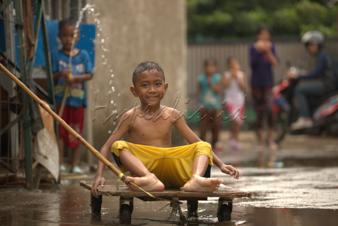 Foto ini memperlihatkan seorang anak laki-laki muda yang sedang menikmati kegembiraan bermain di tengah genangan air hujan. Senyum cerahnya menggambarkan kepuasan dan kesenangan yang tak terhingga. Matanya berbinar dengan cahaya polos, mencerminkan semangat petualangan yang membara di hatinya.<br />
Pakaiannya yang kuning cerah seolah bercahaya di tengah suasana basah, menjadi simbol keceriaan di tengah kesuraman. Ekspresi wajahnya yang penuh harap menggugah hati, mengingatkan kita pada masa kanak-kanak yang penuh keceriaan dan tanpa beban.<br />
Foto ini mengandung pesan yang mendalam tentang kebahagiaan sederhana yang bisa ditemukan di tengah kesulitan. Anak laki-laki itu mengajarkan kita untuk tidak kehilangan semangat dalam menghadapi tantangan hidup. Ia menunjukkan bahwa kebahagiaan bukanlah sesuatu yang harus dicari di tempat jauh, melainkan bisa ditemukan dalam momen-momen sederhana, seperti bermain di genangan air hujan