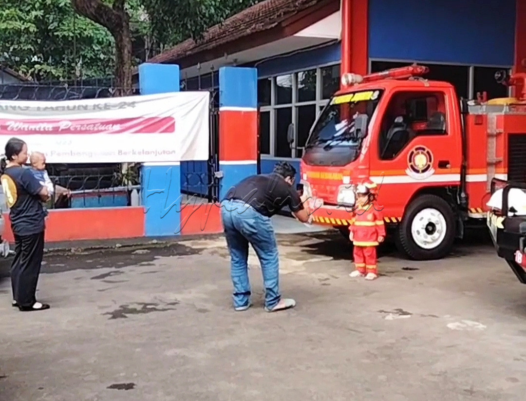 Tampak seorang anak sedang berfoto-foto di depan mobil damkar, dan dia sangat bangga karna dr usia sejak dini sudah mempunyai baju anti api nya