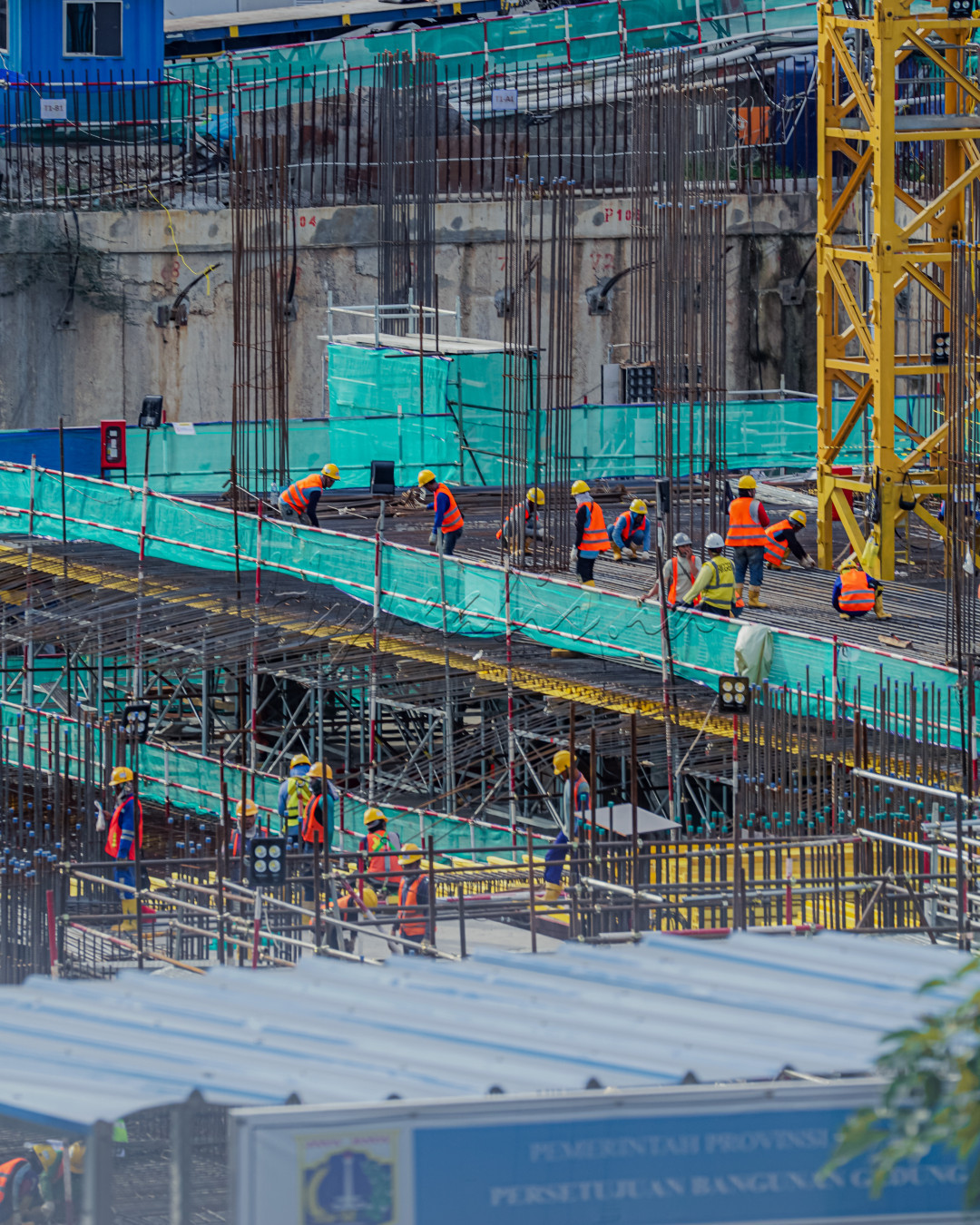 Para pekerja konstruksi yang sedang sibuk membangun infrastruktur. Dengan helm dan rompi keselamatan, mereka bekerja di tengah rangka baja dan material bangunan. Suasana kerja sama terlihat jelas, menggambarkan semangat gotong royong untuk membangun negeri. Ini adalah potret nyata bagaimana kerja keras dan dedikasi menjadi bagian dari upaya menciptakan masa depan yang lebih baik.