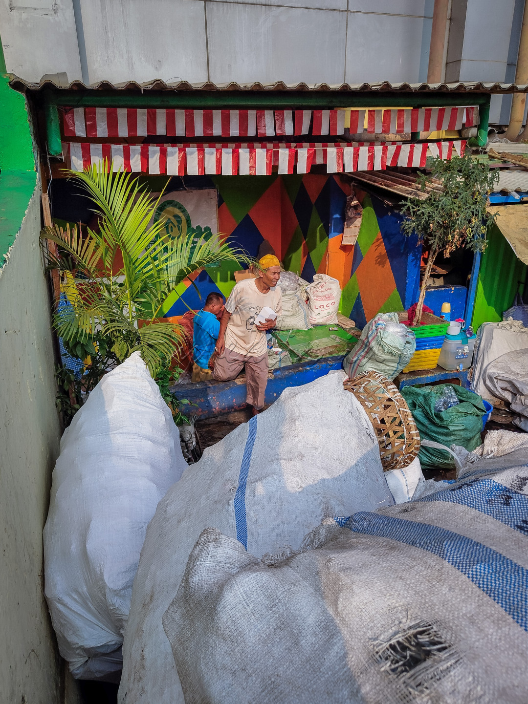 Di sudut kecil kota, seorang pria tua bekerja keras mengumpulkan barang bekas. Dengan latar dinding penuh warna, tempat ini menjadi saksi perjuangan mereka yang jarang terlihat, namun berperan besar dalam menjaga lingkungan dan ekonomi. Ini adalah wujud nyata semangat membangun negeri dari akar rumput.